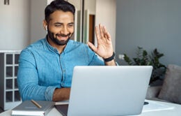 Smiling man wearing wireless earphones waving hand at laptop screen.