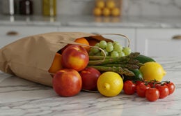 A paper grocery bag overflows with fresh fruits and vegetables on a marble countertop