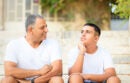 Teenager son and senior father sitting on stairs outdoors at home, talking.