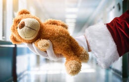 Santa Claus's hand wearing a white glove holding a teddy bear in a hospital corridor.