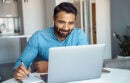 Portrait of young adult indian man wearing earphones looking at laptop computer screen. Motivated student writing notes during online lesson, watching webinar, sitting at work desk