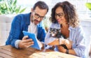 Middle age hispanic couple using touchpad sitting on table with dog at terrace
