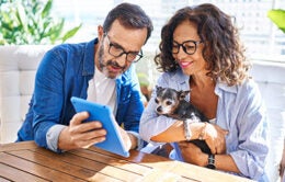 Middle age hispanic couple using touchpad sitting on table with dog at terrace