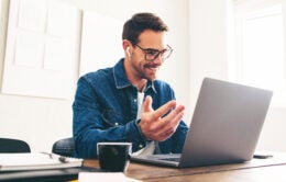 Happy businessman smiling while speaking with his team on a video call. Cheerful businessman having an online meeting on a laptop. Businessman with eyeglasses working remotely in a creative office.