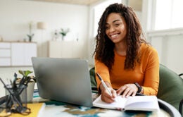 Smiling young African American woman sitting at desk working on laptop and journaling in notebook