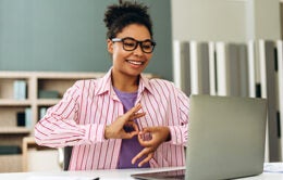 Happy young woman wearing glasses is using sign language while having an online conversation