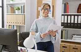 A smiling woman with short blonde hair holding documents stands in a modern office interior.