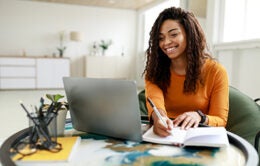 Smiling young African American woman sitting at desk working on laptop taking notes in notebook