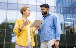 Two business colleagues discuss work using a tablet outside a modern office building, emphasizing collaboration and communication.